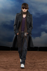 Male model walking a fashion runway in a dark trench coat, brown fringed scarf, sunglasses, and white boots on a sandy surface