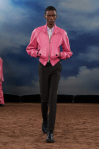 Male model walking a fashion runway in a bright pink bomber jacket, light shirt with a tie, black trousers, and black lace-up boots against a blue cloudy backdrop.