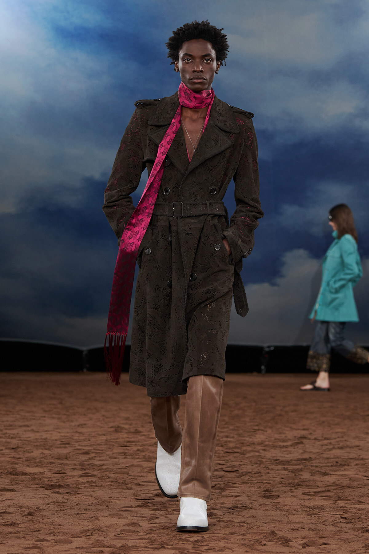 Runway model wearing a dark lace trench coat with a belt, bright pink scarf, and two-tone brown boots against a blue sky backdrop.