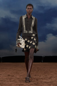 Model walking on a runway wearing a black shirt with lace trim and a circular black-and-cream skirt, carrying a white handbag on the left.