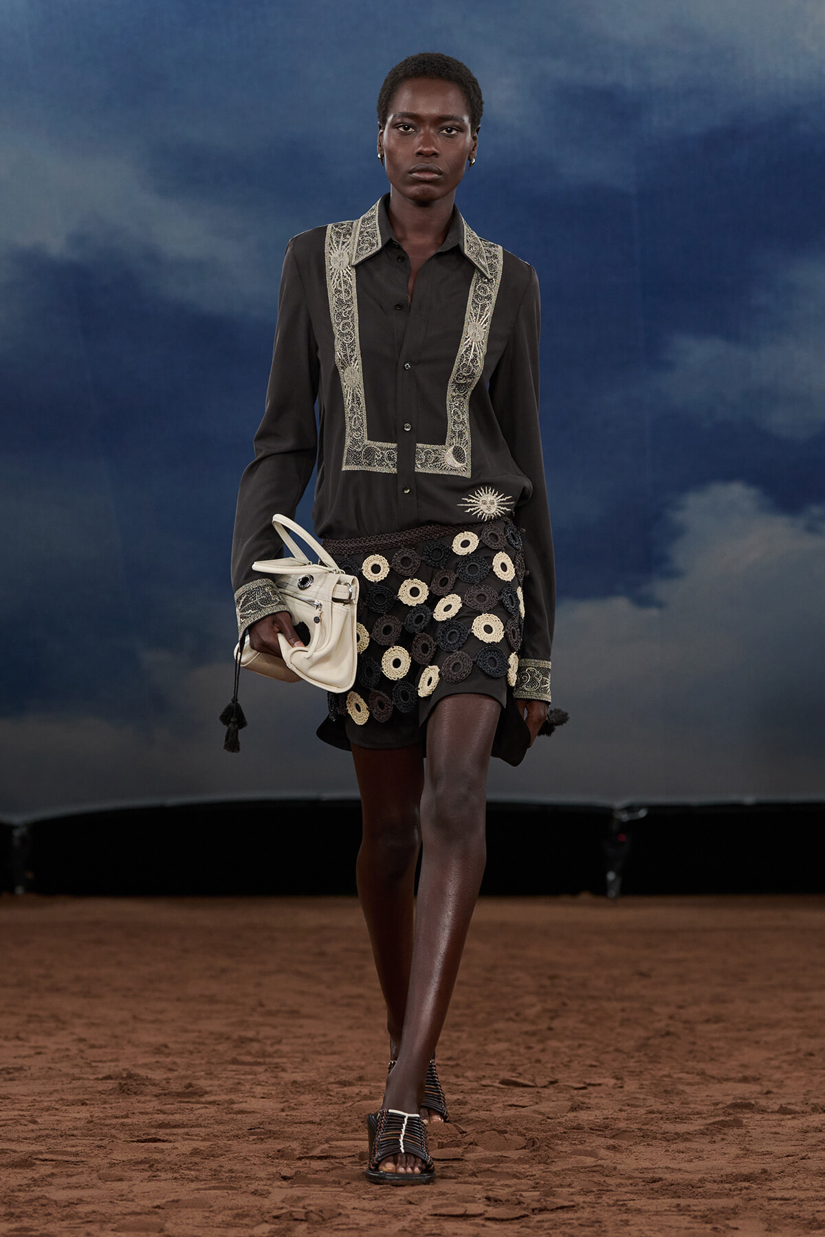 Model walking on a runway wearing a black shirt with lace trim and a circular black-and-cream skirt, carrying a white handbag on the left.