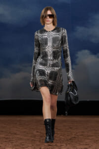 Model walking a fashion show in a silver sequined plaid mini dress, black sunglasses, and black combat boots, carrying a black handbag.