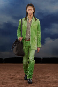 Model walking a runway in a lime green suit with a brown belt and checkered shirt, carrying a dark handbag.