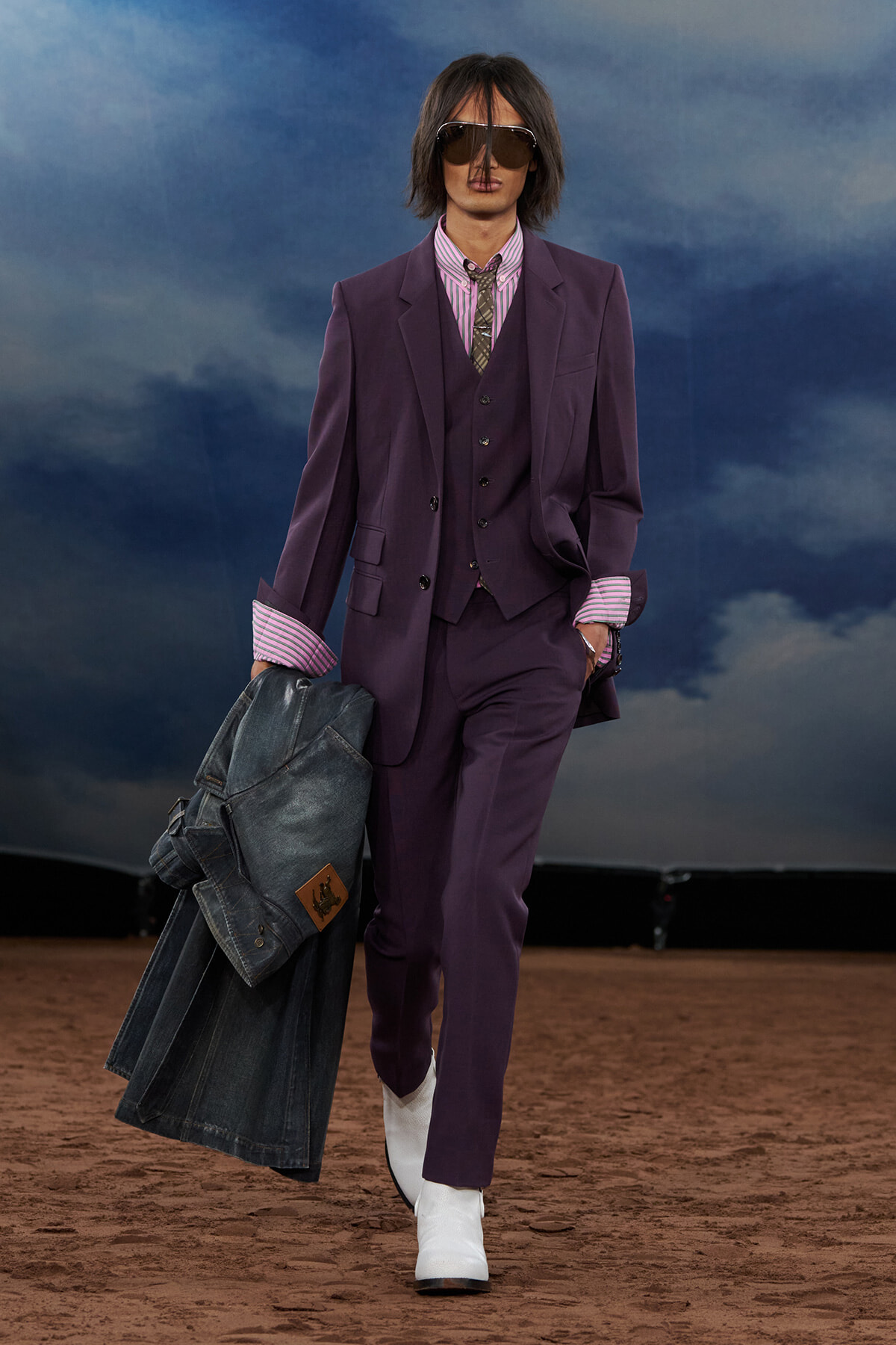 Model walks a fashion runway in a purple suit, striped shirt, and sunglasses, carrying a jean jacket and wearing white ankle boots.