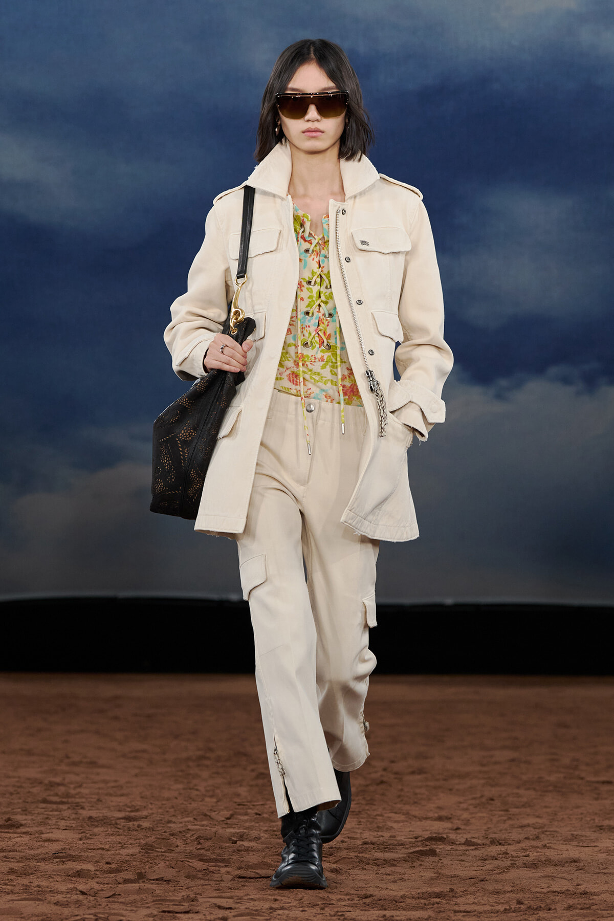 Model walks a runway in a cream utility jacket and cargo pants, floral top, oversized sunglasses, and a black handbag.