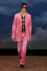 Male model on a runway wearing a pink blazer and matching pink pants with a brown argyle sweater, accessorized with pink sunglasses and black boots.