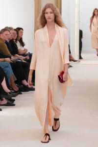 Model walks a peach draped gown with deep V neckline, red clutch in hand, sandals, audience watching on left.