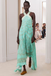 Model walking the runway in a mint-green halter dress with gold embroidery and brown shoes in a bright venue.