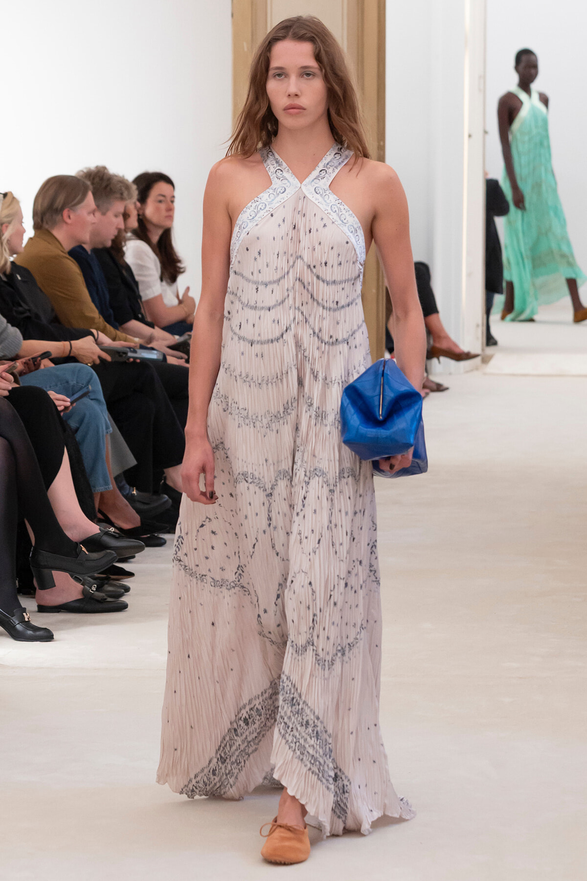 Model walking a runway in a pale, beaded halter gown with blue embroidery, carrying a royal blue clutch.