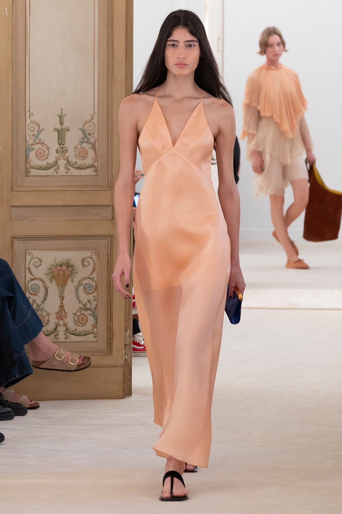 Model walks the runway in a peach satin halter-neck maxi dress, carrying a small blue clutch.