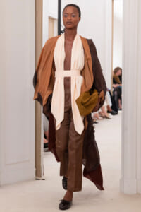 Model walking the runway in a layered tan, cream, and brown draped gown with a wide belt and oversized sleeves.