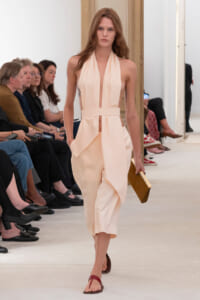 Model in a pale peach halter dress walks the runway, holding a gold clutch while seated guests watch on both sides.