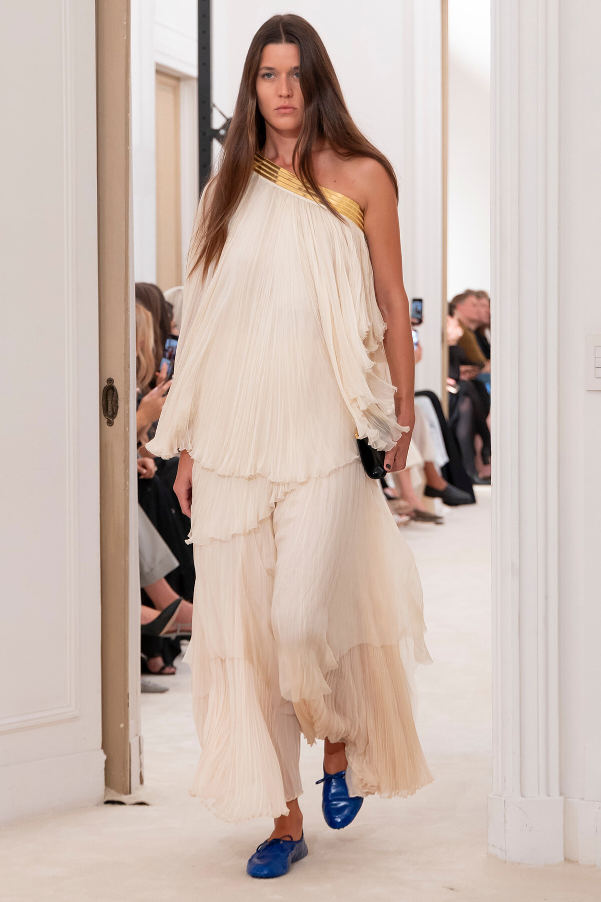 Female model in an off-white, pleated one-shoulder gown with a gold strap, blue shoes, walking down a runway.