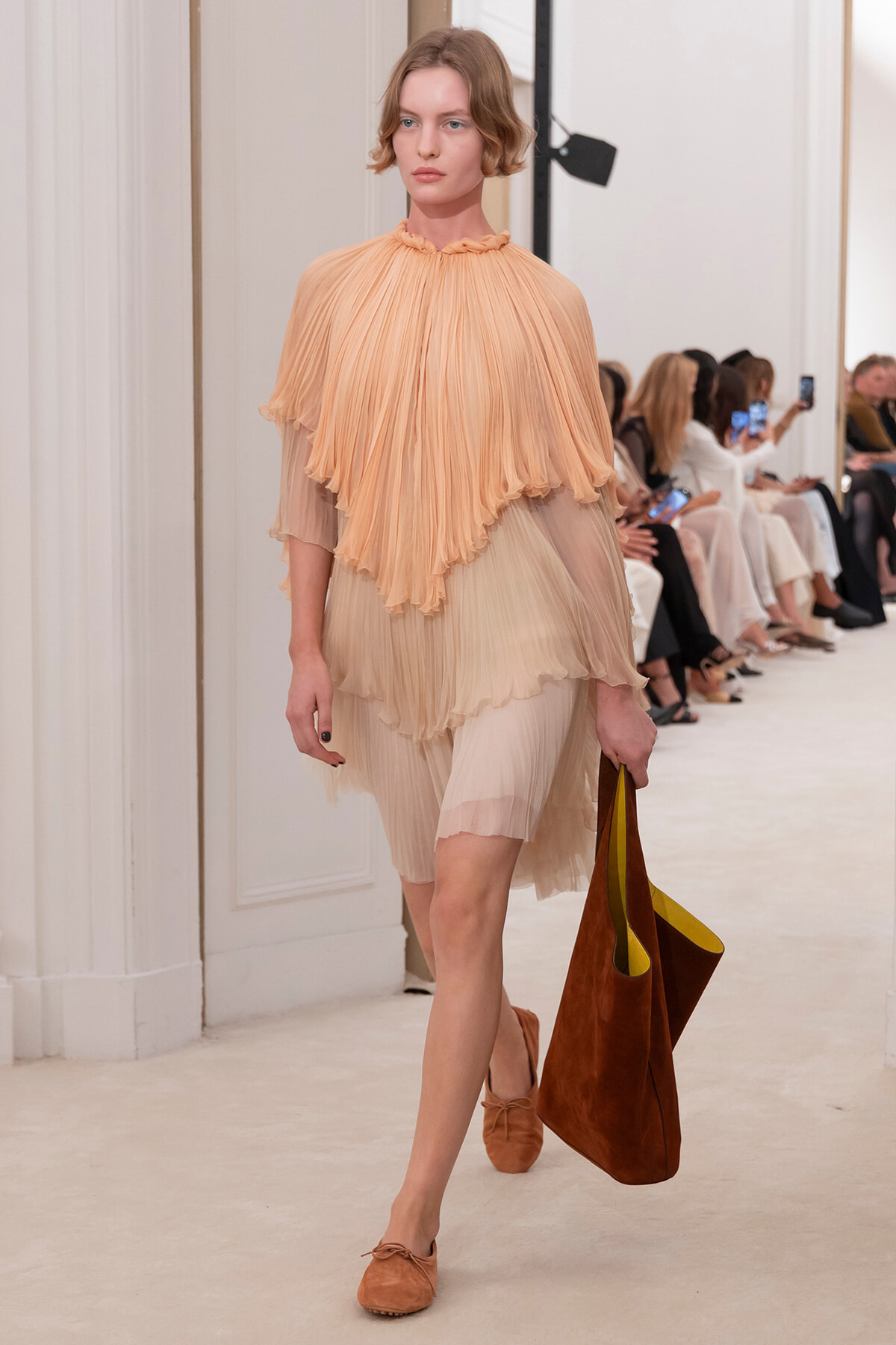 Model walking a runway in a peach layered pleated dress, carrying a large brown tote bag and wearing matching suede flats.