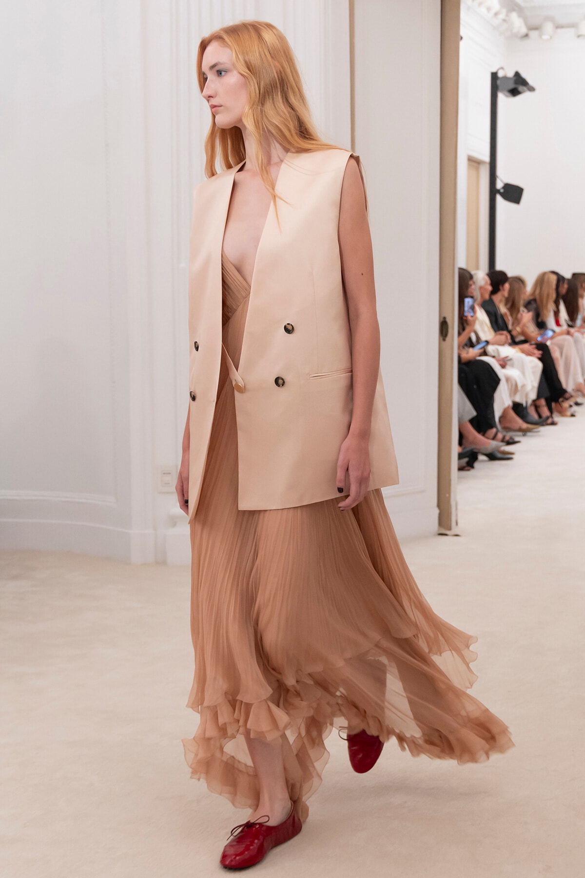 Model walking a fashion show in a peach-toned sleeveless blazer over a sheer tiered gown, with red flats and an audience seated in the background.