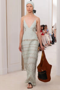 Model walks a runway in a pale mint-green spaghetti-strap dress with lace panels, matching headscarf, and a large brown suede tote with a black front pocket.