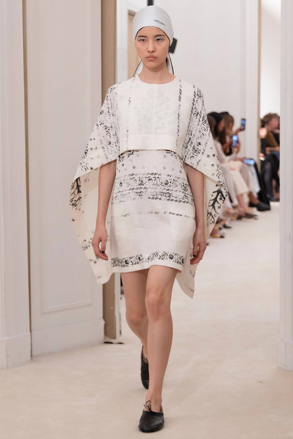 Model walking the runway in a white patterned cape over a matching dress, with a white headscarf, in a fashion show hallway.