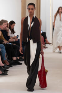 Model walking a fashion show runway wearing a black and white draped outfit with a burgundy vest, carrying a large red textured tote bag to the side.