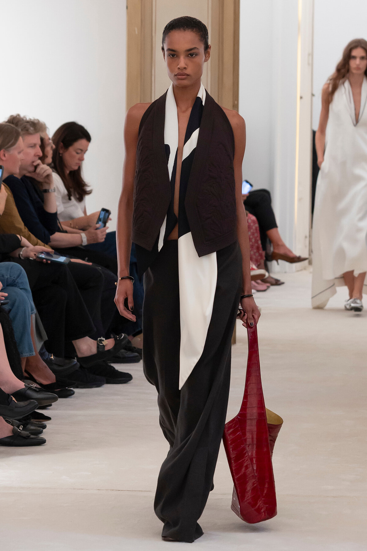 Model walking a fashion show runway wearing a black and white draped outfit with a burgundy vest, carrying a large red textured tote bag to the side.