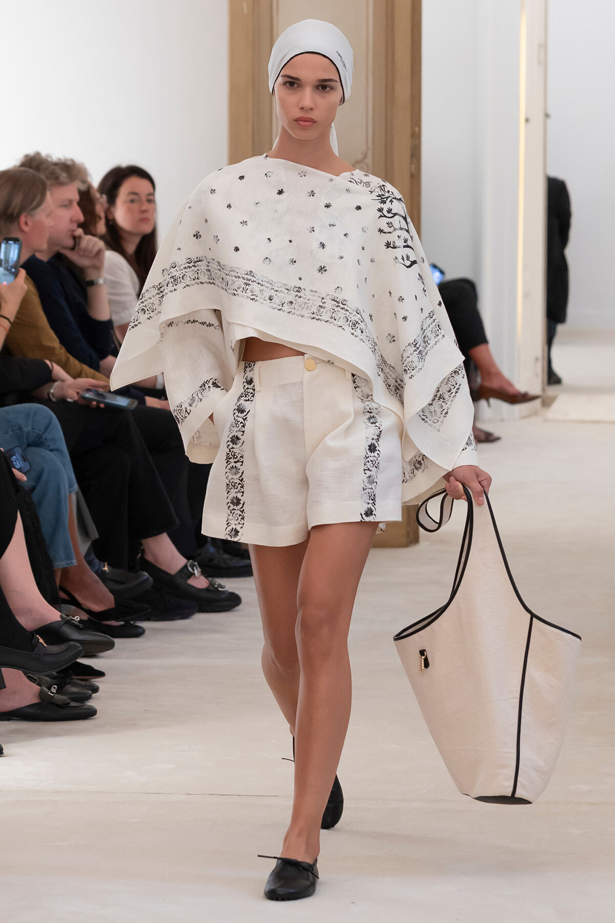 Model walking a runway in a white embroidered poncho and matching shorts, white headscarf, black flats, and a large beige tote with black trim.