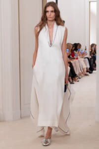 Model walking a runway in a white sleeveless gown with a deep V neckline and black piping along the edges.",