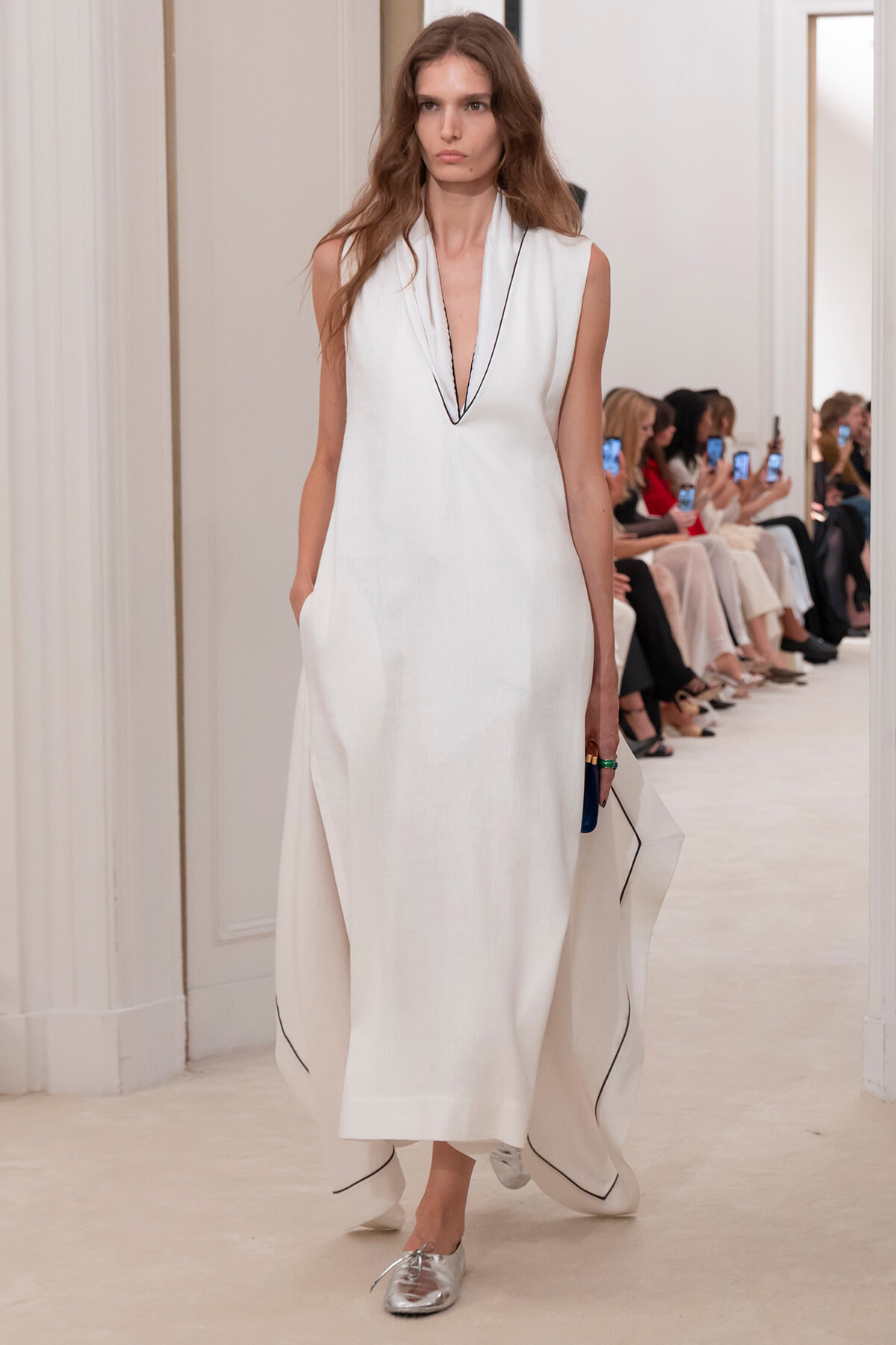 Model walking a runway in a white sleeveless gown with a deep V neckline and black piping along the edges.",