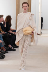 Model walking a runway in a cream textured wrap over crochet wide-leg pants, holding a beige hat.