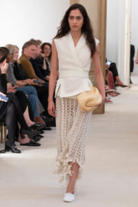 Model walks the runway in a white textured wrap top and long crochet skirt, holding a tan woven hat.