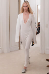 Model walking a fashion show in a white deep-V jumpsuit with long, wide sleeves and silver shoes.