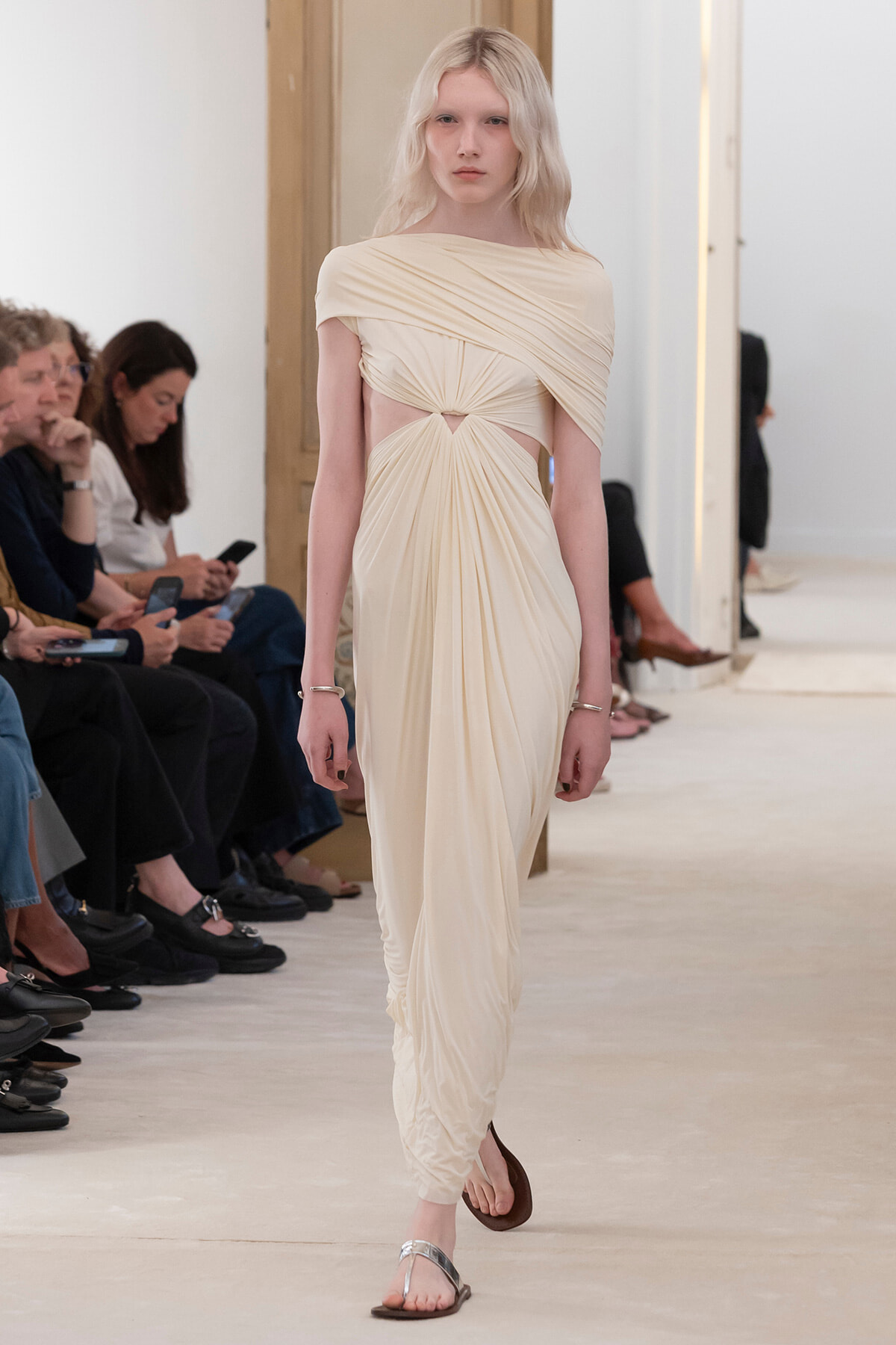 Model walking the runway in a cream draped gown with a twisted waist on a fashion show catwalk; seated audience along the sides.