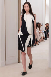 Model walking a fashion show in a black-and-white draped dress with scarf-like detailing, audience seated along the runway in the background.