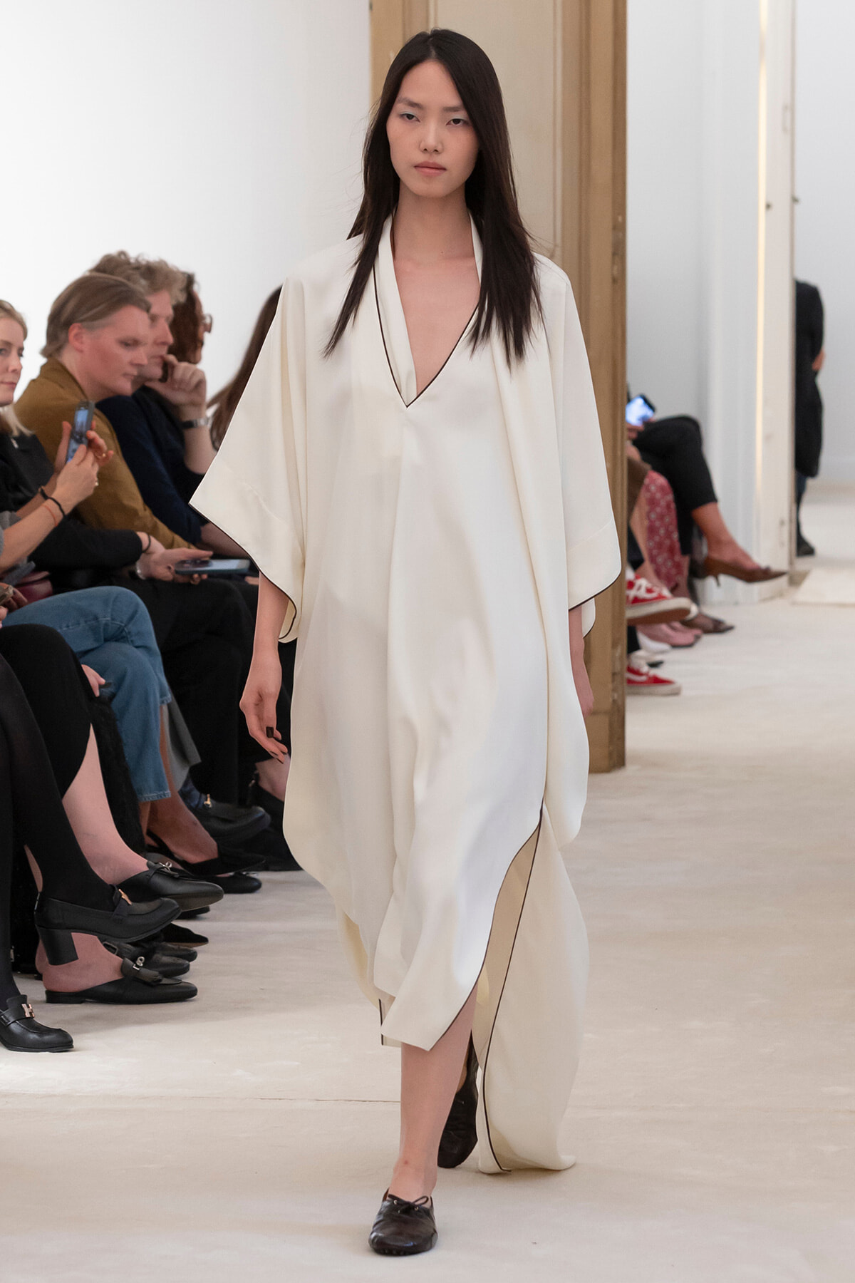 Model walking a runway in a cream, draped robe with a deep V-neck and black trim; audience seated along the catwalk.