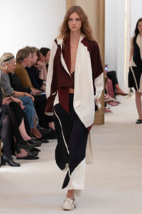 Model on a runway wearing a burgundy and cream color-block coat with a deep neckline over wide-leg pants; audience seated along the catwalk.