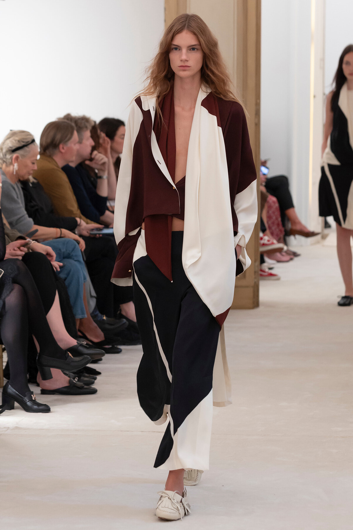 Model on a runway wearing a burgundy and cream color-block coat with a deep neckline over wide-leg pants; audience seated along the catwalk.
