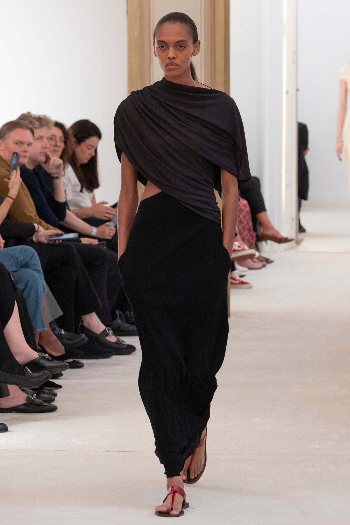 Model walking a runway in a black draped top and long black skirt with red sandals, audience seated along the catwalk.