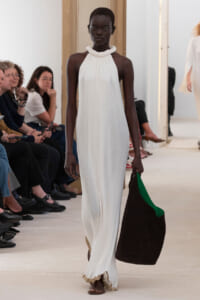 Runway scene: dark-skinned model in a white halter gown walks forward, carrying a large brown bag with a green interior.