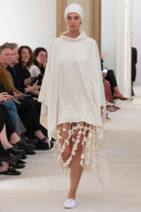 Model on a fashion runway wearing an all-white outfit: flowing cloak with a hood, head wrap, and a fringed floral-hem skirt.