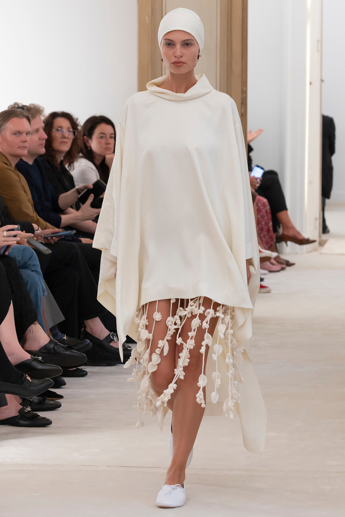 Model on a fashion runway wearing an all-white outfit: flowing cloak with a hood, head wrap, and a fringed floral-hem skirt.