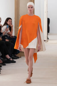 Model walking the runway in an orange and white color-block cape dress, wearing a white headscarf and tan sandals.