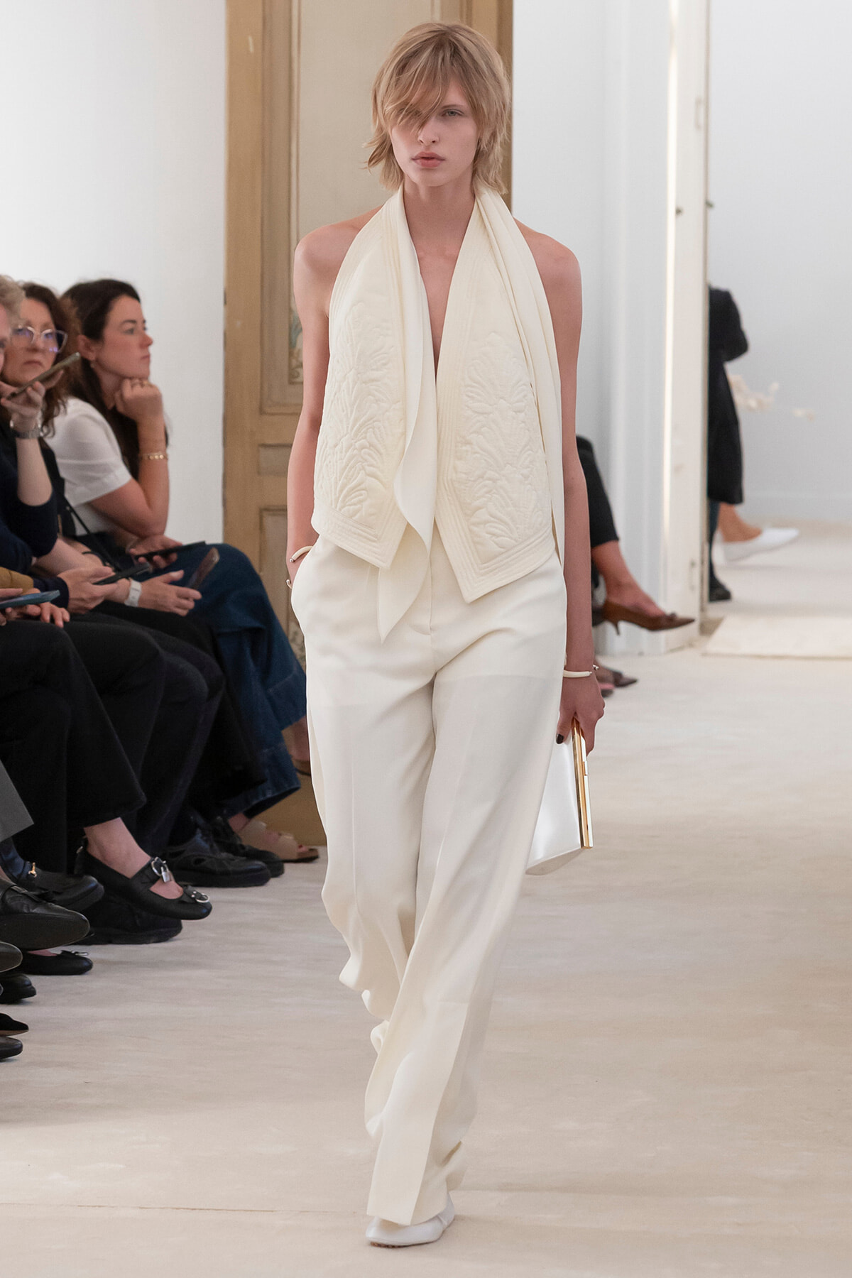 Model walking the runway in an all-white ensemble: draped scarf-like top over high-waisted wide-leg pants, white boots, and a white clutch. -Alternative 1: Female model with short blonde hair in ivory sleeveless top and matching trousers, carrying a white clutch. -Alternative 2: Runway model in white, textured scarf detail over a plunging neckline top and wide-leg pants, audience watching.