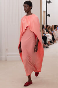 Model walks the runway in a flowing peach-pink draped cape dress with a gathered neckline and red flats.