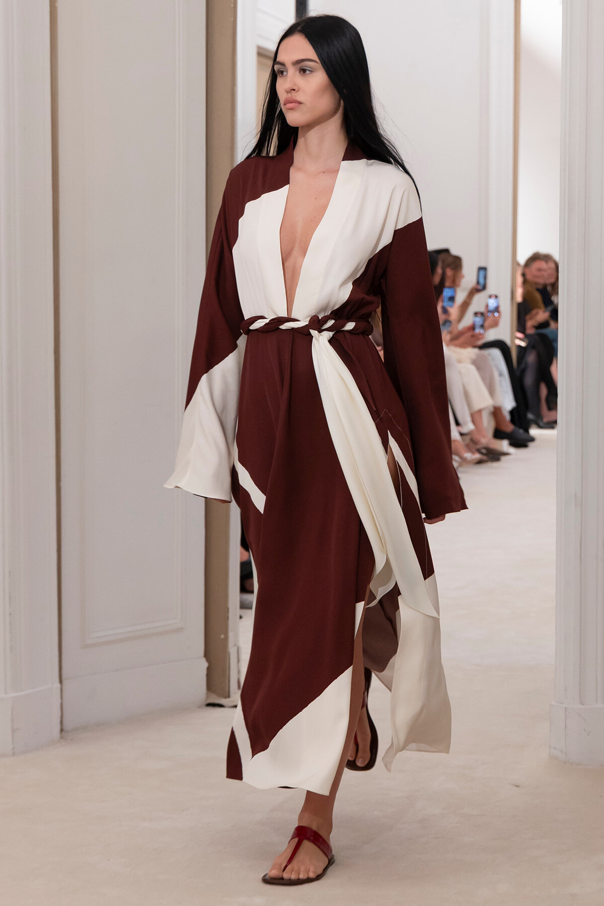 Model walking a runway in a brown and cream color-block wrap dress with a braided belt and deep V neckline.