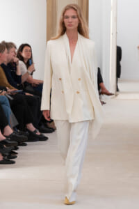 Model walks the runway in a white double-breasted suit with a cape, gold buttons, and pale yellow shoes; audience seated nearby.