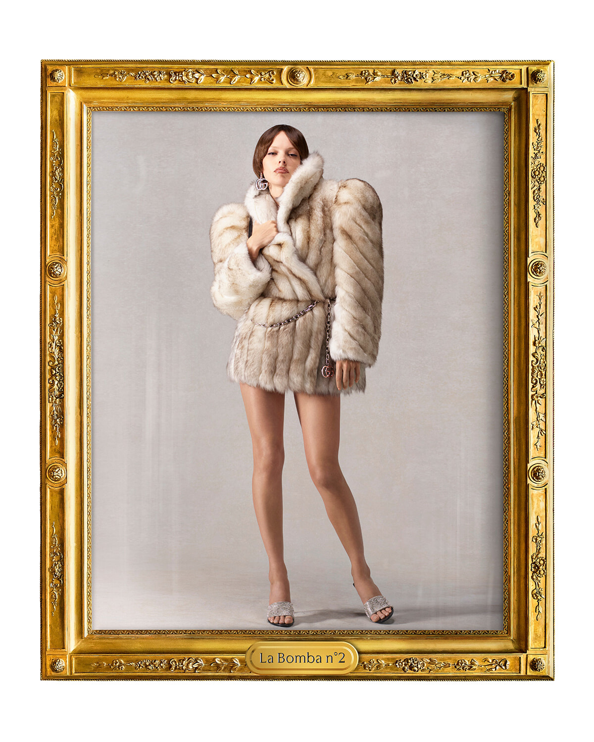 Model wearing a white fur coat with bare legs and silver sandals stands inside an ornate gold frame labeled La Bomba n°2, mid-pose for a high-fashion portrait.