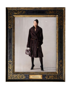 Fashion photo of a woman in a dark fur coat with a gold chain belt and handbag, standing against a neutral backdrop inside an ornate framed portrait frame labeled Milanesa.