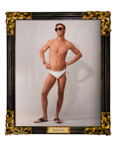 Man in white swim trunks posing with hands on hips, wearing sunglasses, inside an ornate black-and-gold frame labeled Bastardo.