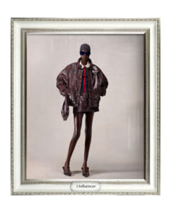 Fashion portrait of a tall model wearing a metallic brown oversized jacket, striped top, cap, sunglasses, and heels, standing confidently with a handbag on a beige backdrop.