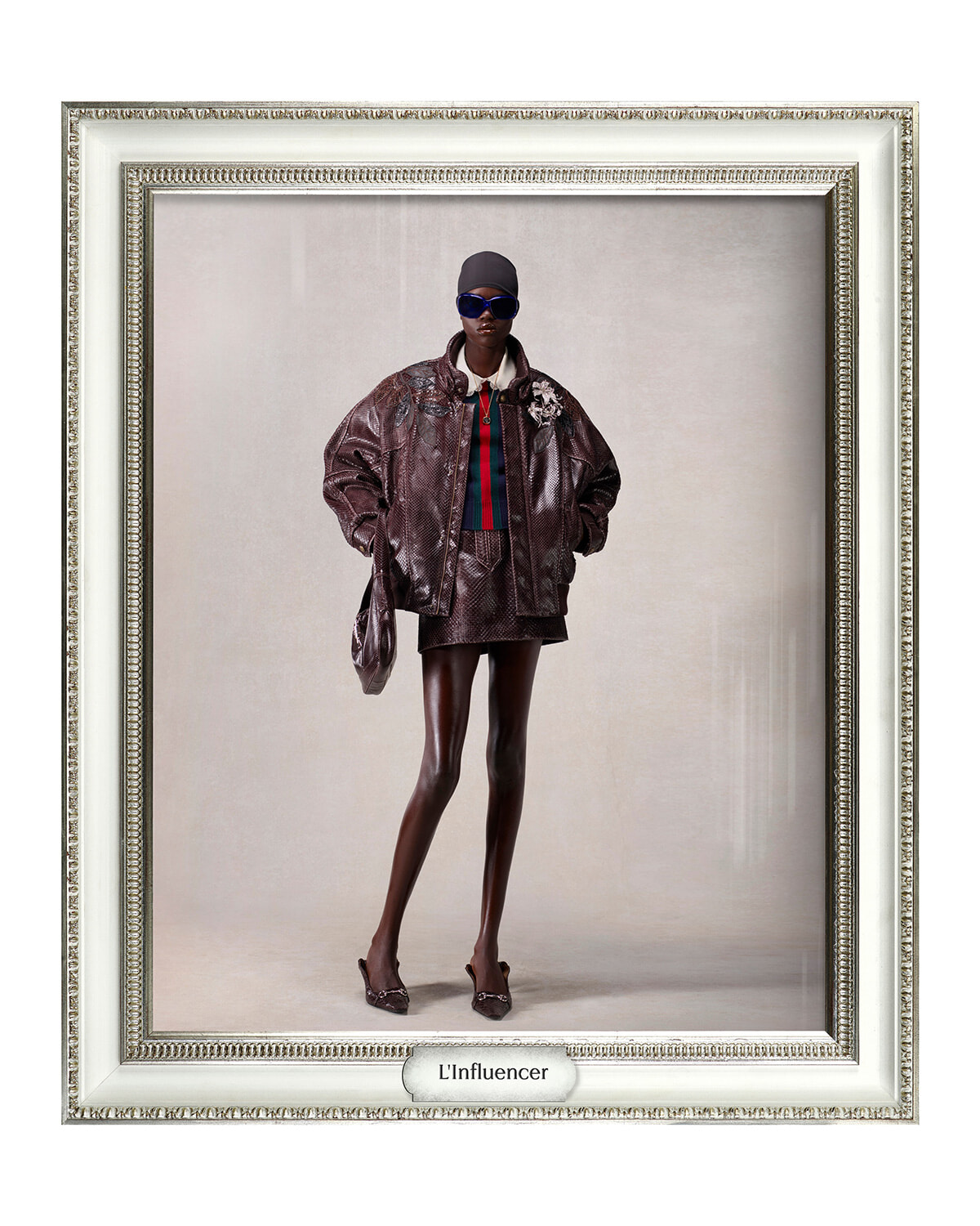 Fashion portrait of a tall model wearing a metallic brown oversized jacket, striped top, cap, sunglasses, and heels, standing confidently with a handbag on a beige backdrop.