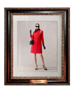 Elegant portrait of a woman in a red coat, black gloves, headscarf and sunglasses, holding a black handbag, framed like gallery art with a bronze plaque reading 'Incazzata'