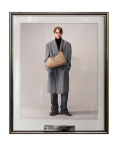 Model wearing a gray plaid overcoat over a black turtleneck, holding a beige patterned shoulder bag in front of a plain backdrop (framed portrait).
